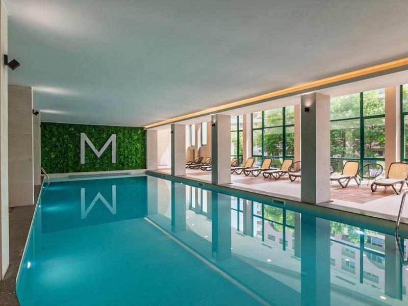 Piscina cubierta con tumbonas y grandes ventanas con vista a árboles verdes