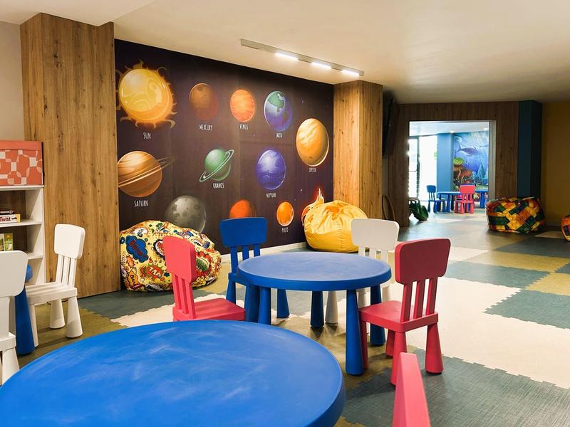 Sala de juegos infantil colorida con mesas, sillas y decoración de planetas en la pared en un hotel.
