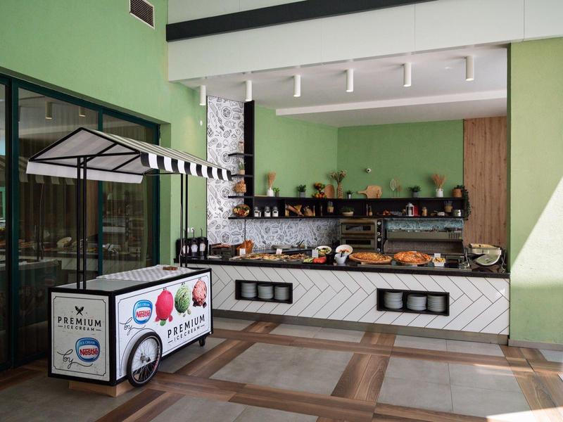 Zona de desayuno moderna con buffet, pasteles y carrito de helados en un hotel luminoso.