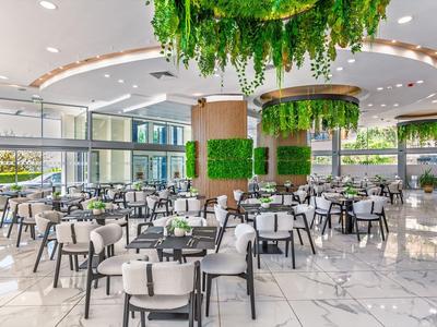 Moderní hotelová restaurace s bílými stoly, židlemi a bohatou zelenou rostlinnou výzdobou.