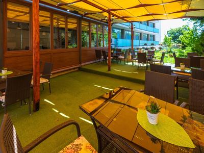 Zastřešená terasa s dřevěnými stoly a židlemi, obklopená trávou a stromy u hotelu.