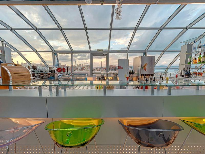 Moderne Bar mit transparentem Dach, bunten Stühlen und Regalen voller Flaschen und Gläser.