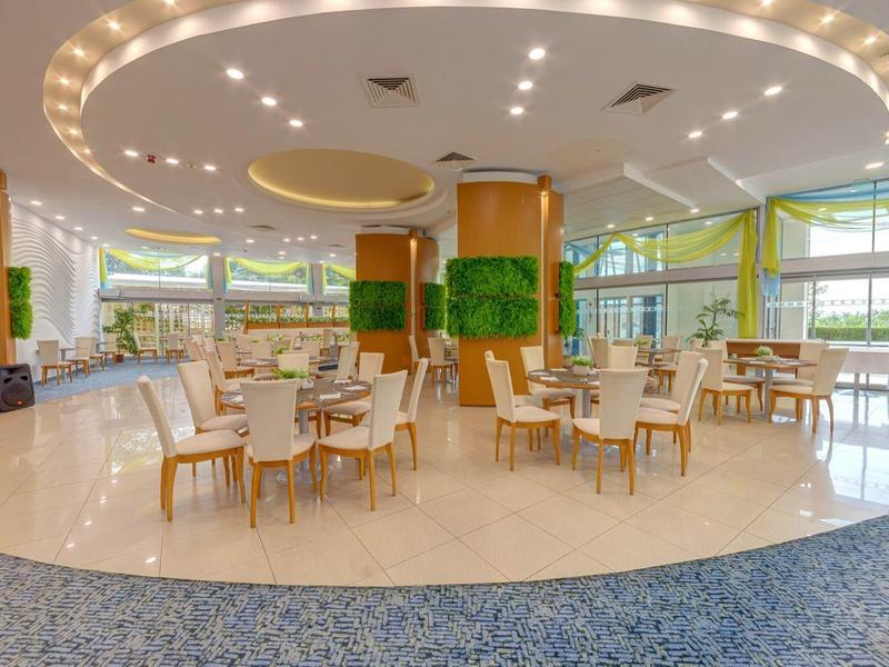 Großer, heller Speisesaal mit runden Tischen, beigen Stühlen und deckenhohen Fenstern.