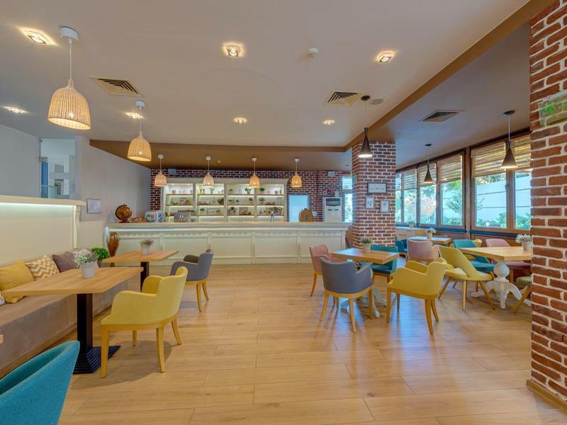 Moderner Gastraum mit Holzboden, bunten Stühlen, langer Sitzbank und großen Fenstern.