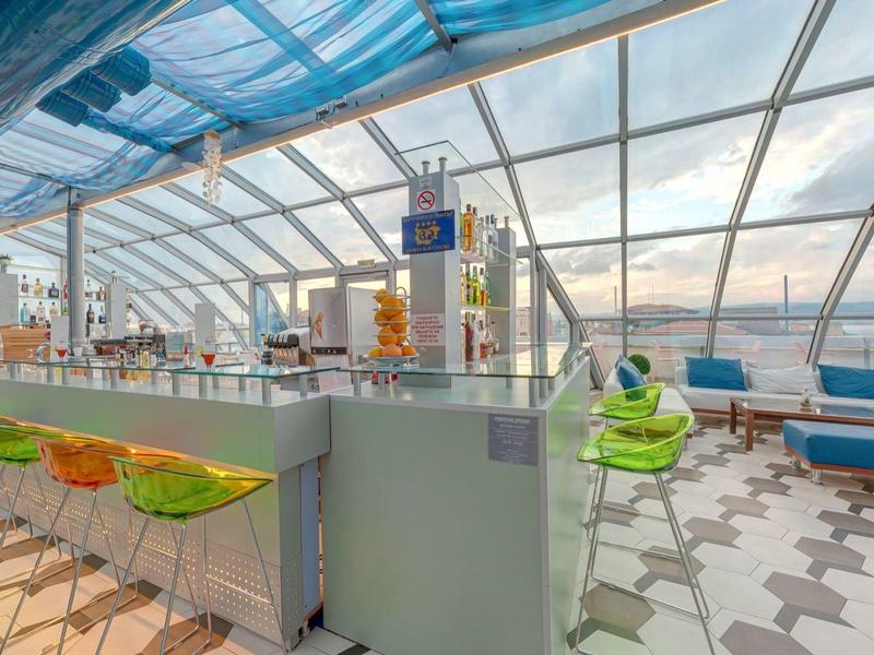 Moderne Dachterrasse mit Glasüberdachung, farbigen Stühlen und Blick auf Hafen und Himmel.