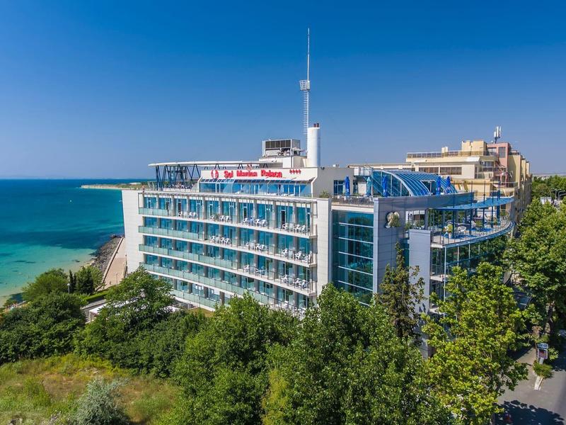 Modernes Hotel am Meer mit blauer Fassade und umliegendem Grün bei klarem Himmel.