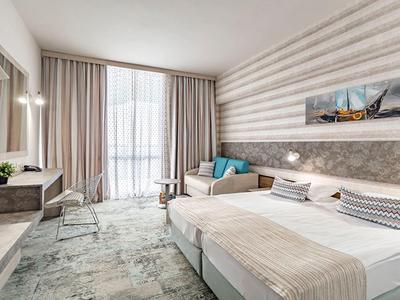 Helles Hotelzimmer mit Doppelbett, Sesseln, Teppichboden und großen Fenstern mit Vorhängen.