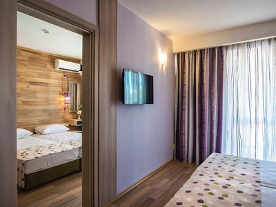 Modernes Hotelzimmer mit Blick auf ein separates Schlafzimmer, Wandfernseher und große Fenster mit Vorhängen.