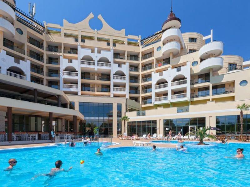 Großes Hotel mit weißen Balkonen und blauen Pool, Leute schwimmen bei sonnigem Himmel.