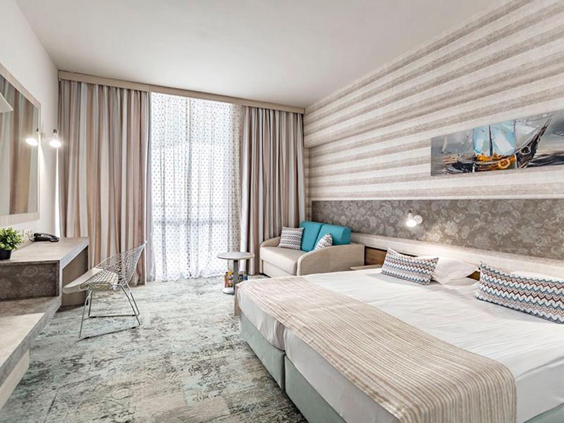 Helles Hotelzimmer mit Doppelbett, Sesseln, Teppichboden und großen Fenstern mit Vorhängen.