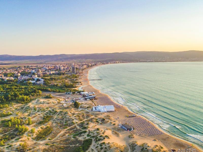 Langer Sandstrand mit Dünen, dahinter grünes Waldgebiet und Stadt, ruhiges blaues Meer bei Sonnenuntergang.