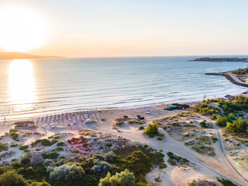 Sonnenaufgang am Sandstrand mit Dünen und Sonnenschirmen, dahinter ruhiges Meer und Himmel.