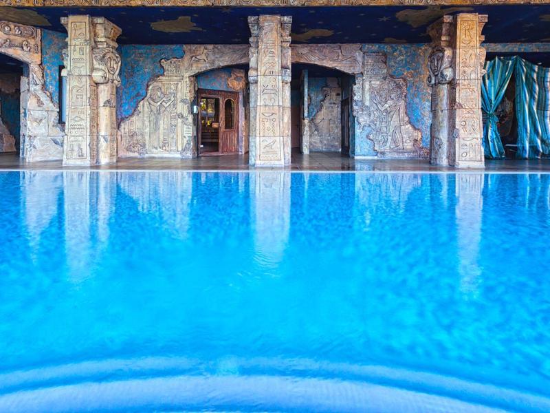 Blaues Wasserbecken mit reflektierender Oberfläche vor einer Wand mit türkisfarbenem Vorhang und Säulen.