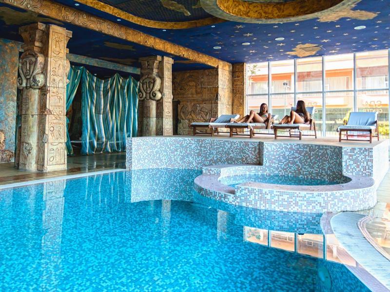 Innen-Schwimmbad mit blauem Wasser, Rundbecken, Säulen und großer Fensterfront bei Tageslicht.