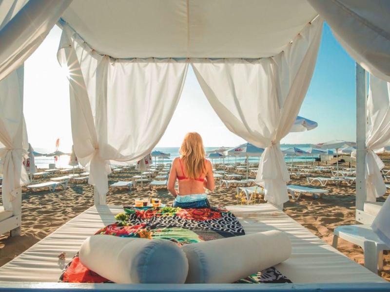 Eine Frau sitzt auf einem weißen Daybed mit Himmelbett am sonnigen Strand mit Lounge-Möbeln.