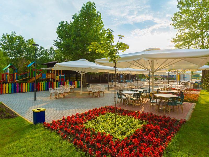 Freiluftrestaurant mit buntem Kinderspielplatz, Sonnenschirmen und Blumenbeet bei Abendlicht.