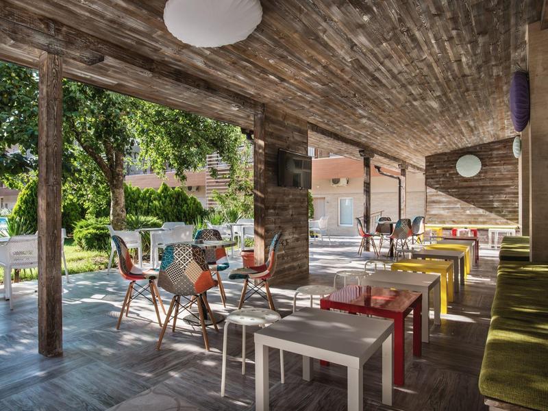 Überdachte Terrasse mit bunten Tischen und Stühlen, Holzbalkendecke und Blick in grünen Garten.