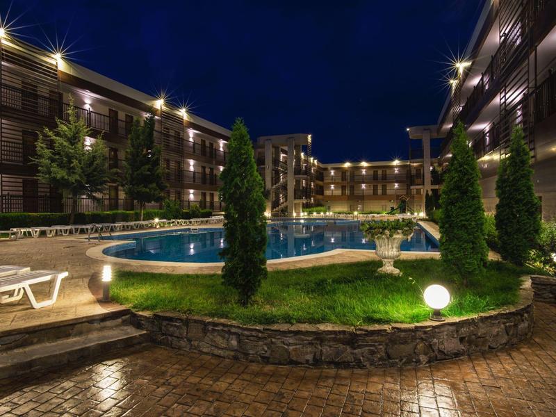 Beleuchteter runder Pool mit umgebender Wiese, Sitzgelegenheiten und Wohngebäuden bei Nacht.