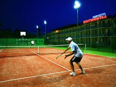 Tennisspieler auf rotem Sandplatz bei Nacht, beleuchtet von Flutlicht, Hotel im Hintergrund.
