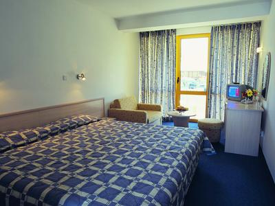 Einfaches Hotelzimmer mit großem Bett, blauem Teppich und Balkon mit Blick nach draußen.