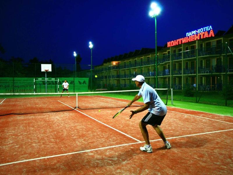 Tennisspieler auf rotem Sandplatz bei Nacht, beleuchtet von Flutlicht, Hotel im Hintergrund.