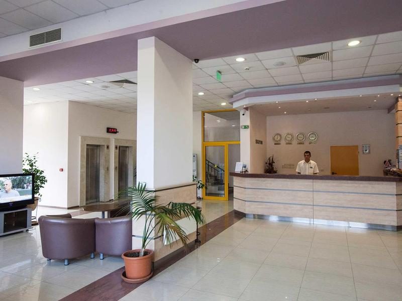 Helle Hotellobby mit Empfangstresen, Sitzgruppe aus violetten Sesseln und grünen Zimmerpflanzen.