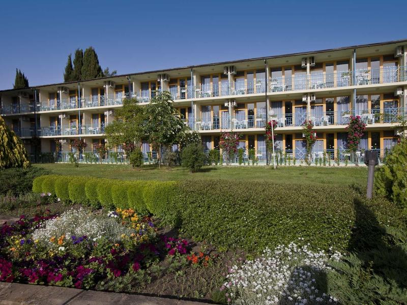Mehrstöckiges Gebäude mit Balkon und gepflegtem Garten mit Blumen und Sträuchern im Vordergrund.