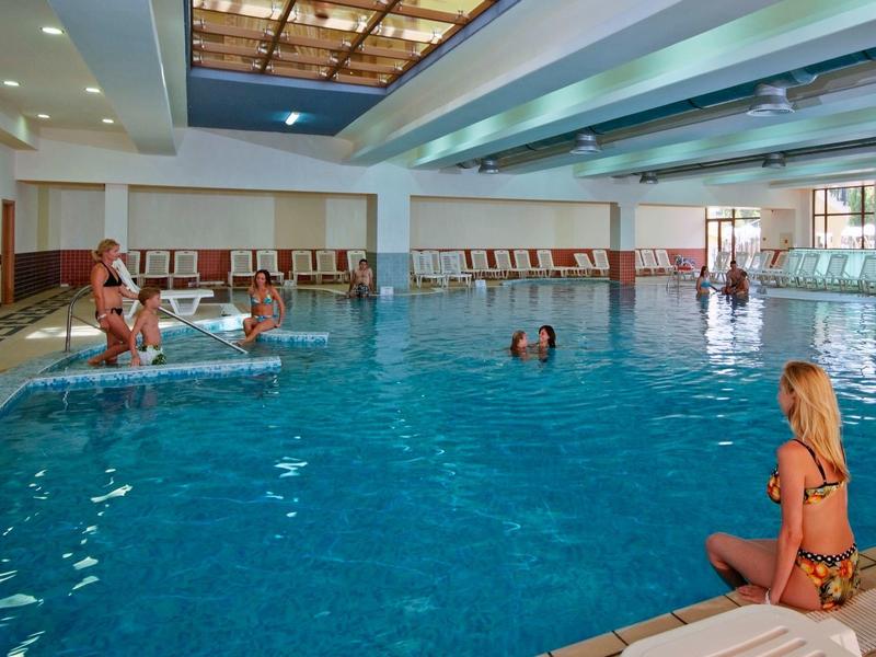 Innenraum-Schwimmbad mit mehreren Menschen, klar blaues Wasser und Liegestühle am Rand.