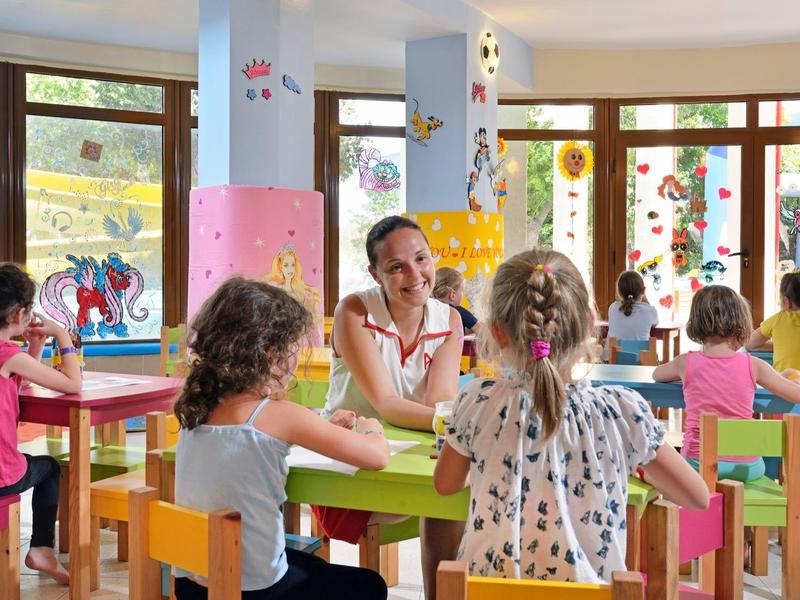 Kinder sitzen an kleinen Tischen in einem farbenfrohen Klassenraum mit großen Fenstern.