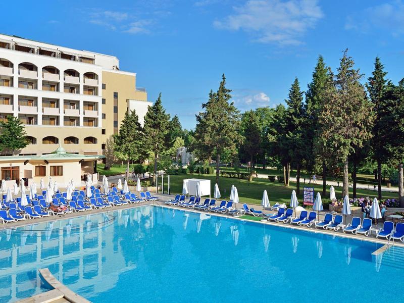 Großer Hotelpool mit Liegestühlen und Bäumen im Hintergrund bei sonnigem Wetter.