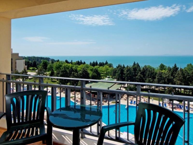 Balkon mit zwei schwarzen Stühlen, kleinem Tisch, Blick auf Swimmingpool, Bäume und Meer unter blauem Himmel.