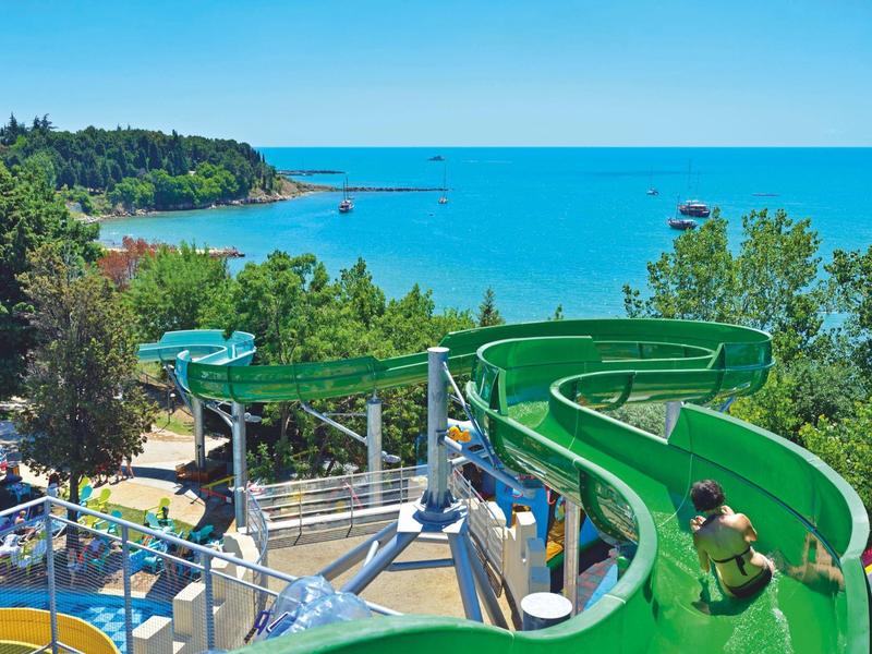 Menschen rutschen eine grüne Wasserrutsche mit Aussicht auf das Meer bei sonnigem Wetter.