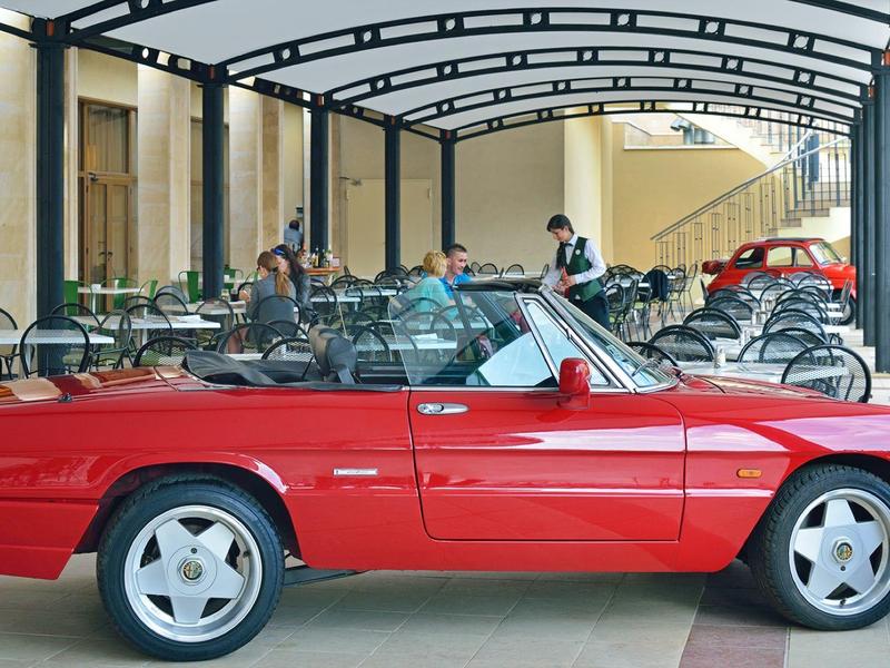 Rotes Cabrio vor einem Hotel mit Sitzgelegenheiten unter einem überdachten Bereich.