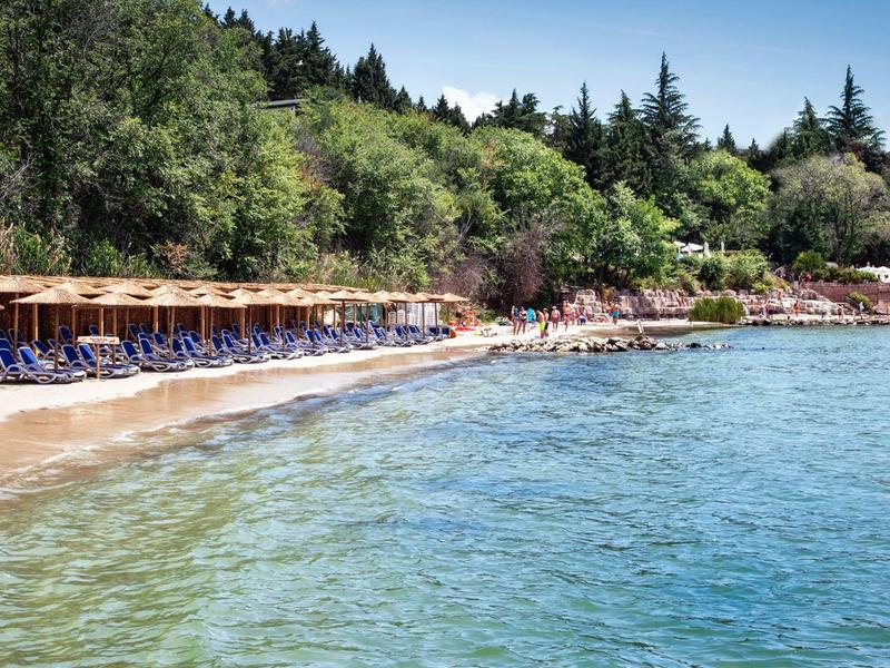 Klarer Strand mit Liegestühlen und Sonnenschirmen neben bewaldetem Hügel.