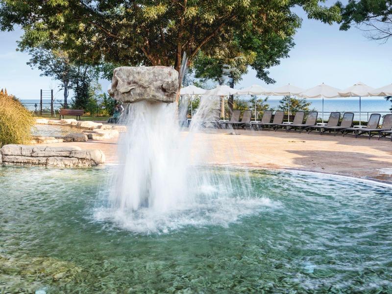 Wasserfontäne in einem Teich mit Liegestühlen und Blick auf das Meer im Hintergrund.