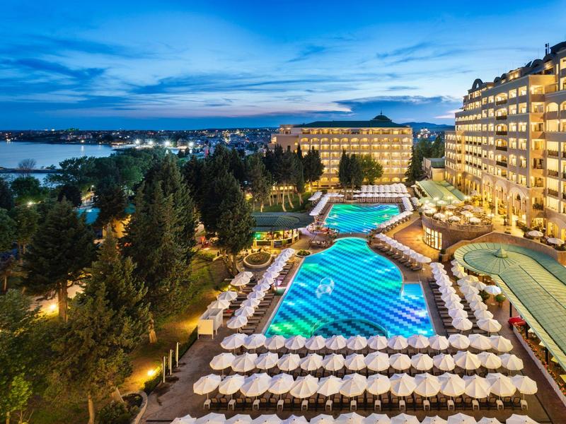 Luxuriöses Hotel mit beleuchtetem Pool und hunderten Sonnenschirmen am Abend mit Blick auf Wasser.
