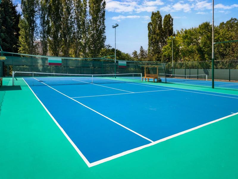 Blauer Tennisplatz mit weißen Linien, grünem Rand und Bäumen im Hintergrund unter blauem Himmel.