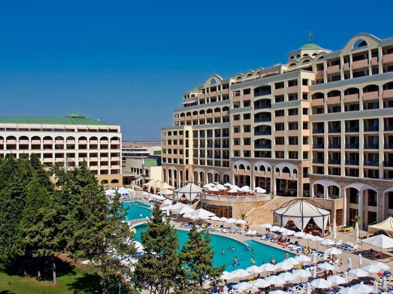 Luxushotel mit großem Pool, Sonnenschirmen und grünen Bäumen unter klarem blauem Himmel.