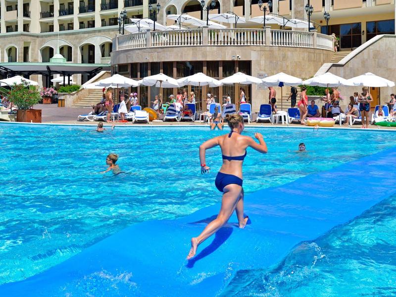Frau springt in einen Pool vor einem Hotel mit Liegestühlen und Gästen.