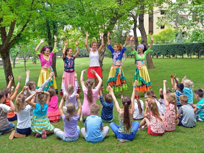 Kinder sitzen im Kreis auf einer Wiese, während andere tanzen, umgeben von Bäumen im Park.