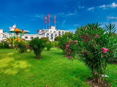 Grüner Hotelgarten mit blühenden Sträuchern und Spielplatz unter blauem Himmel.