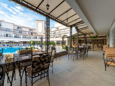 Überdachte Terrasse mit Tischen und Stühlen neben einem Hotelpool unter blauem Himmel.