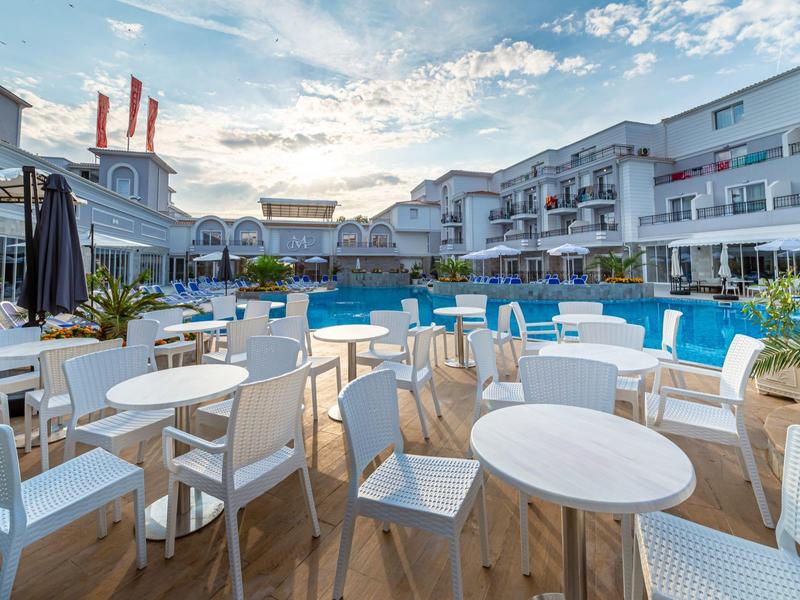 Außenbereich eines Hotels mit weißem Mobiliar, Pool und Sonnenschirmen unter blauem Himmel.