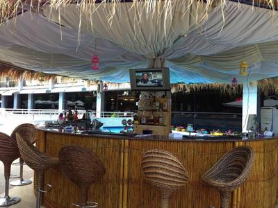 Bar all'aperto con tetto di paglia, sgabelli in rattan e scaffali con liquori.