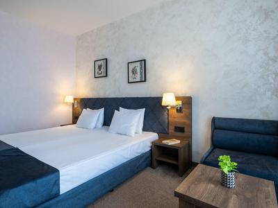 Modern ingericht hotelkamer met tweepersoonsbed, nachtkastje en zithoek met bank en tafel.