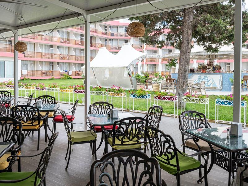 Overdekt buitenterras met metalen stoelen en groene kussens naast een hoteltuin.