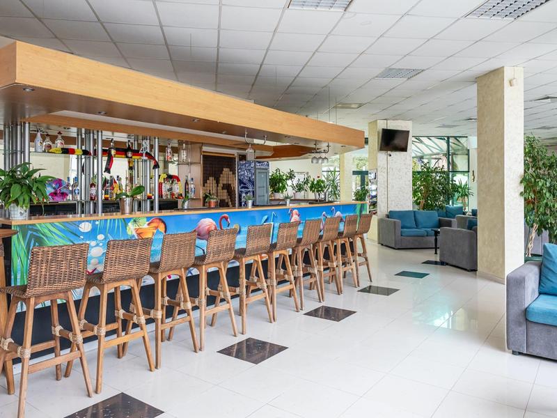 Lichte hotelbar met blauwe krukken, houten meubels en groene planten bij zithoek.