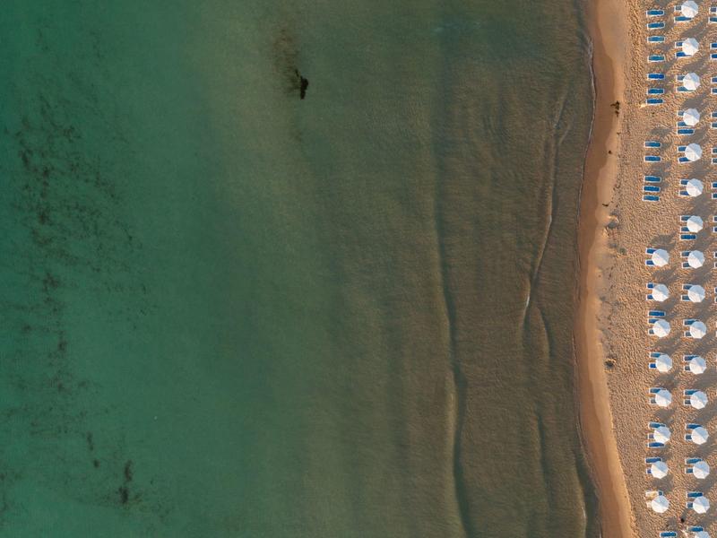 Luftaufnahme eines Strandes mit Reihen von Sonnenschirmen und klarem blau-grünem Wasser.