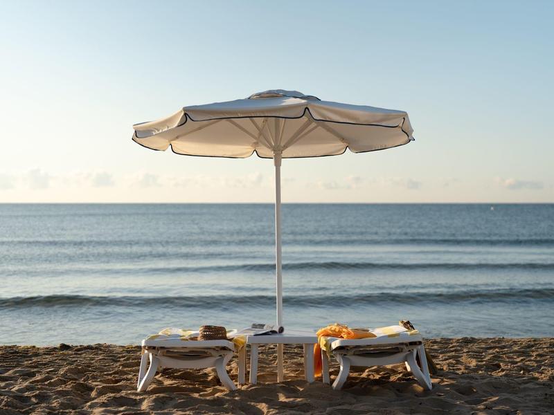 Zwei Liegestühle unter einem Sonnenschirm am Strand mit Meer im Hintergrund.