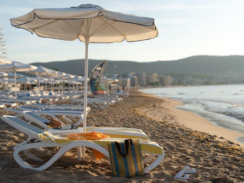 Leerer Liegestuhl und Sonnenschirm am Sandstrand mit Meer und Hügeln im Hintergrund.